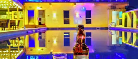 Uma garrafa de cerveja Budweiser e um copo sobre uma piscina reluzente, com edifícios iluminados ao fundo.