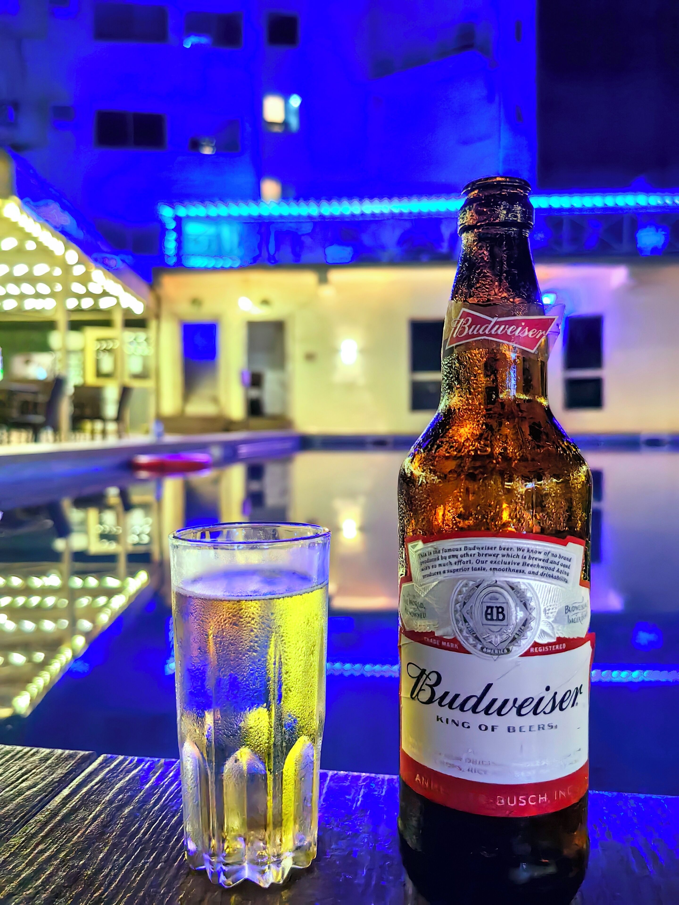 A Budweiser beer bottle and a glass of beer on a table by a poolside with blue lighting.