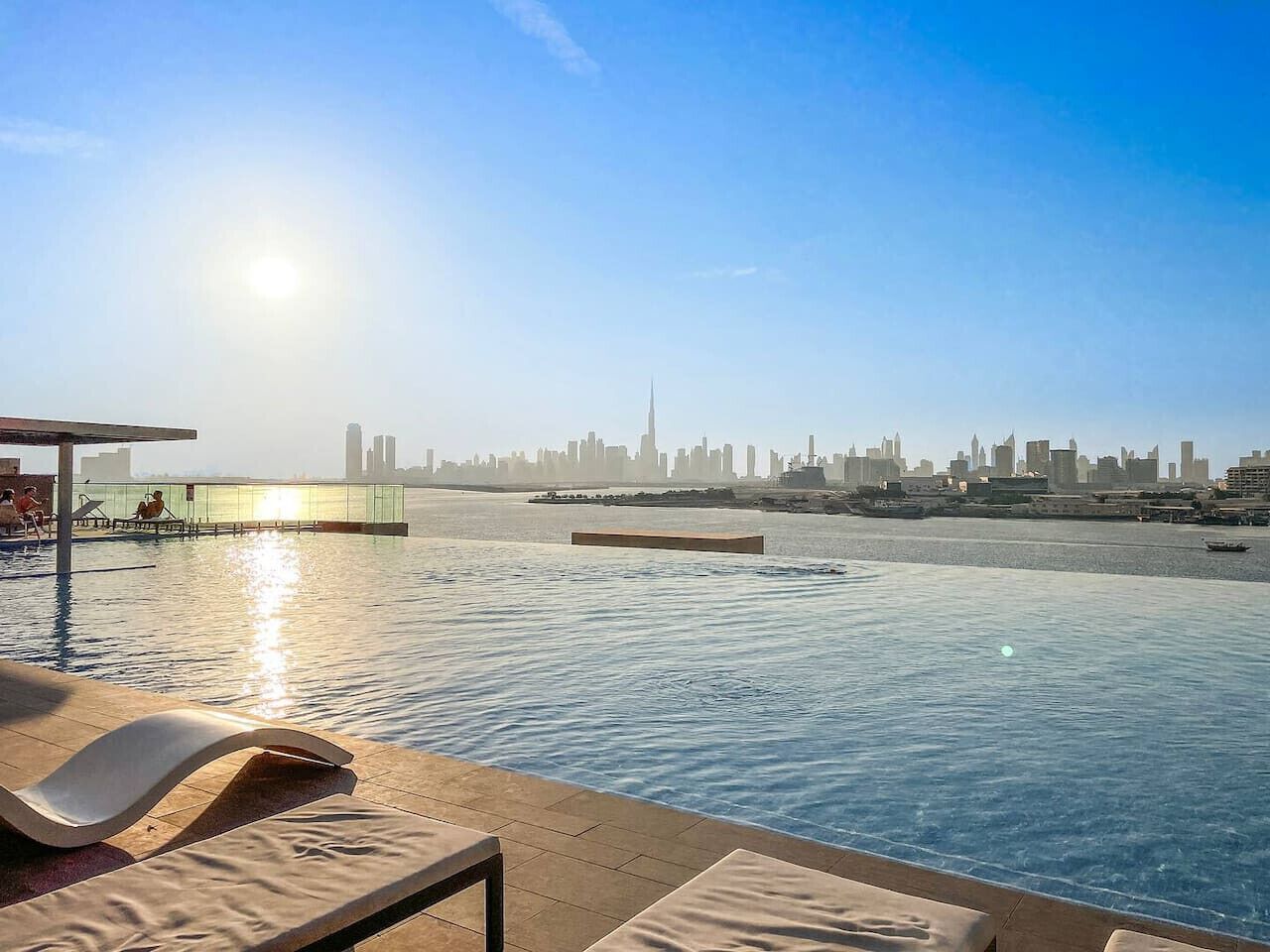 An infinity pool with a city skyline view, lounge chairs, and a sunlit sky.