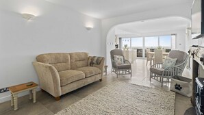 A spacious living area with a beige sofa, two armchairs, a small wooden table, and a large window offering a view of the outdoors.