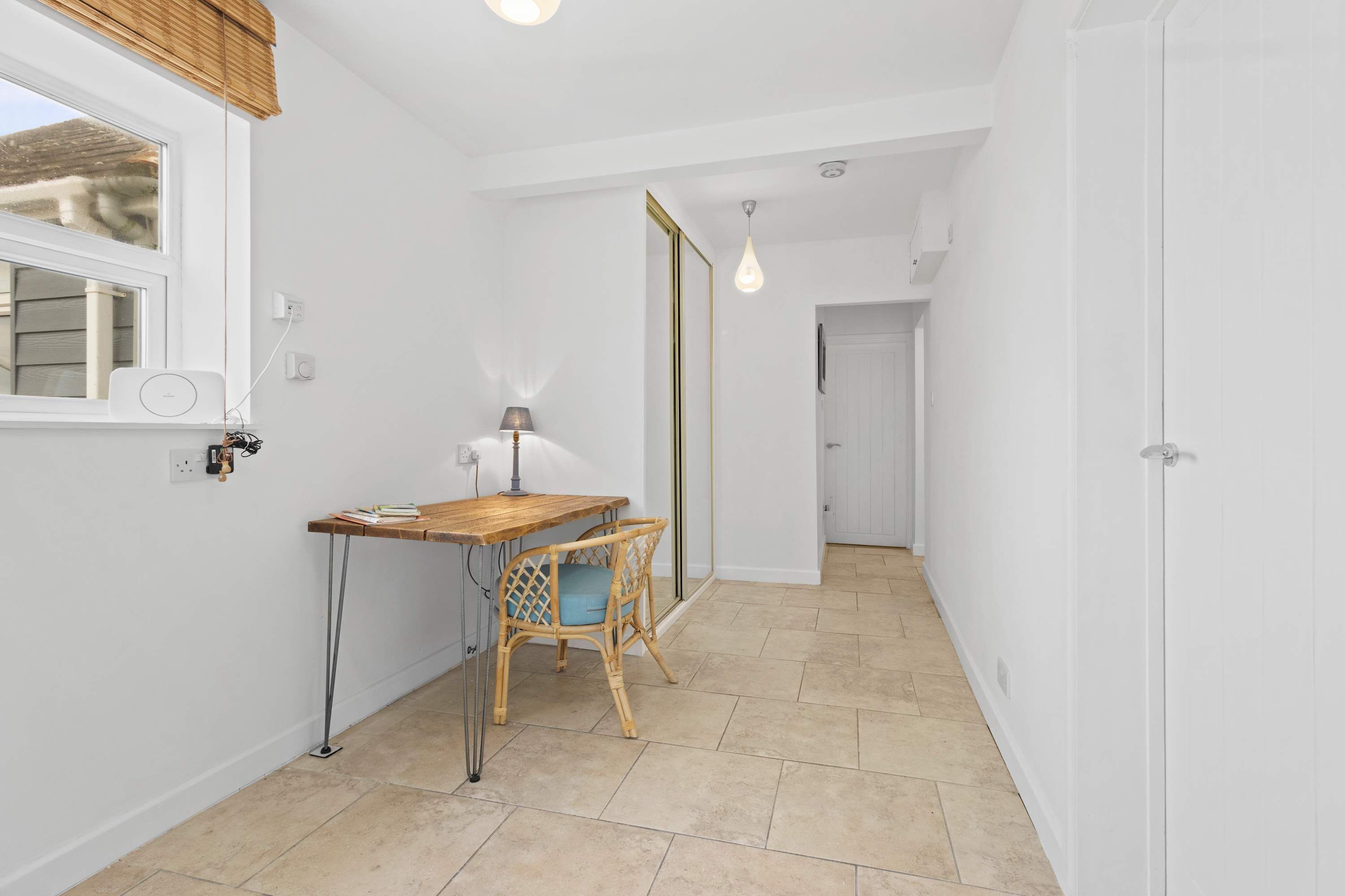 A clean, white-walled room with a wooden desk and chair, a mirror, and a hanging light fixture.