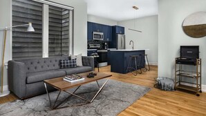 A modern living room with a grey sofa, a wooden coffee table, and a kitchen area with blue cabinets and a stainless steel refrigerator. - The Frankford Getaway (Philadelphia)