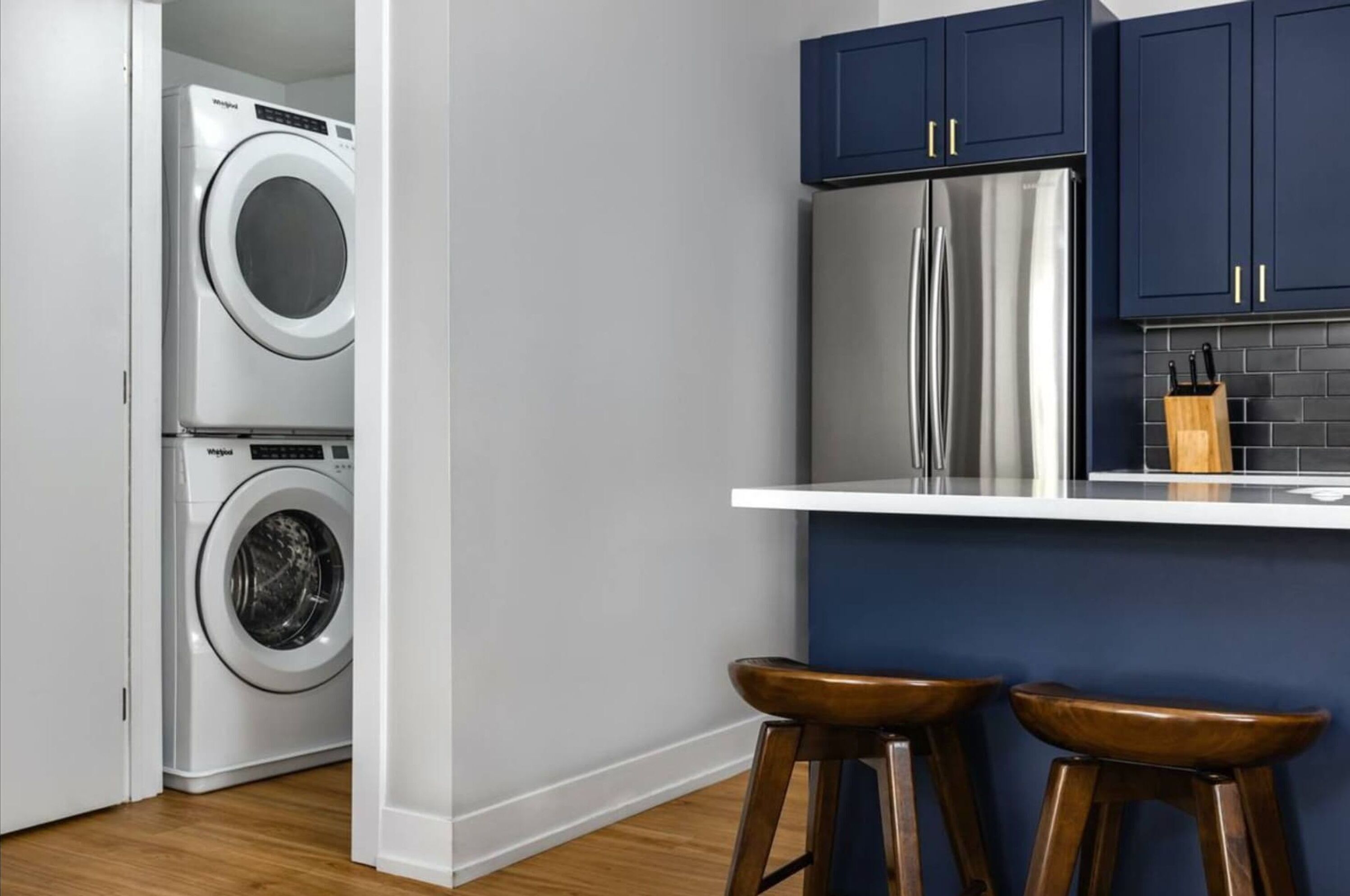A compact kitchen with a stainless steel refrigerator, a washing machine, and two wooden stools.