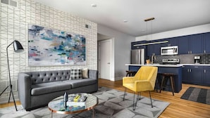 A modern living room with a grey sofa, a yellow armchair, and a coffee table. A large abstract painting adorns the wall, and the room features a geometric patterned rug. - Experience Philly in Style (Philadelphia)