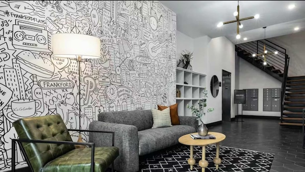 A modern living room with a patterned wall, a grey sofa, a green armchair, a round coffee table, and a staircase in the background. - Philly s Coolest Cozy (Philadelphia)