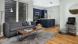 Un salon moderne avec un canapé gris, une table basse en bois et un coin cuisine doté de meubles bleus et d’un réfrigérateur en acier inoxydable.