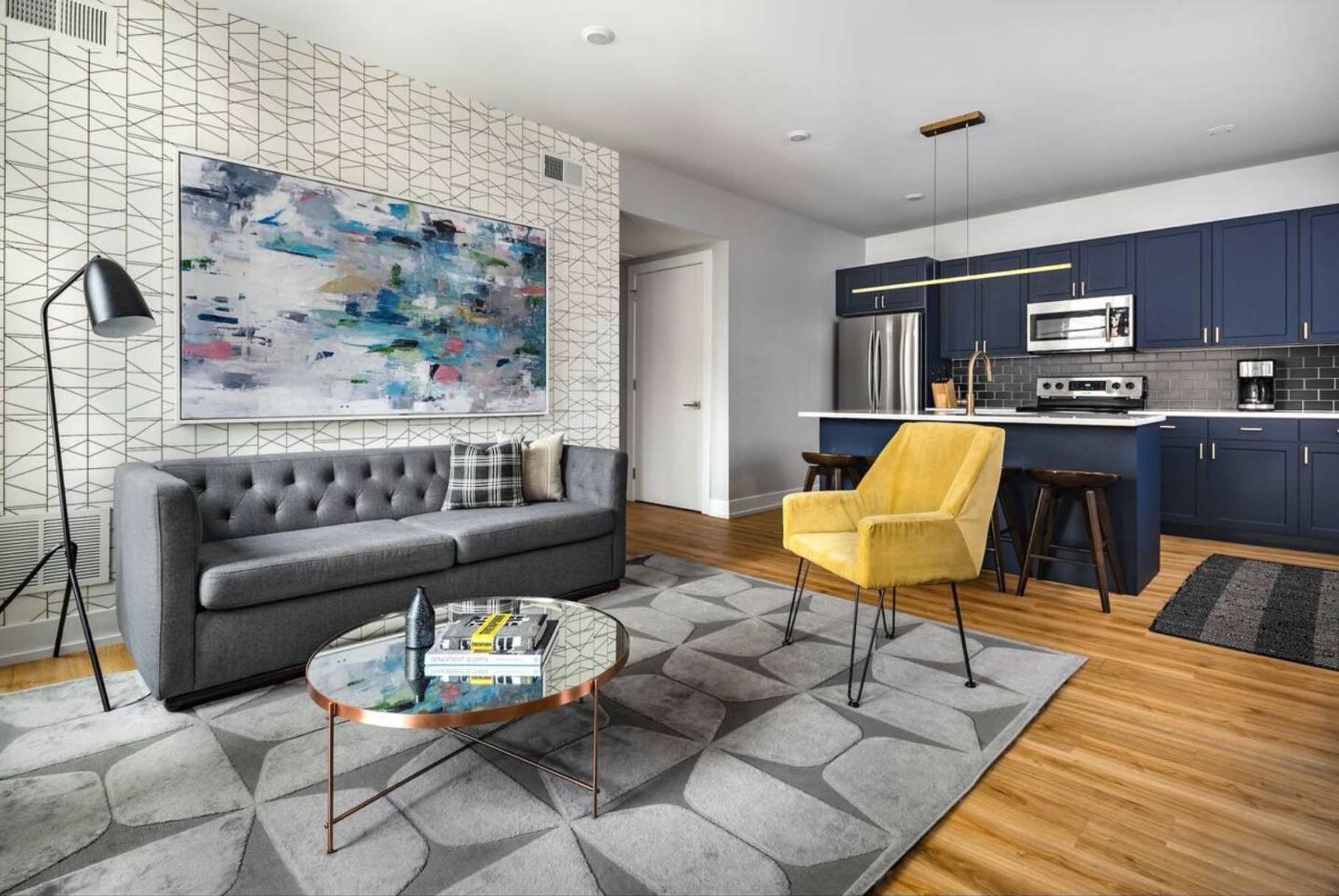 A modern living room with a grey sofa, a yellow armchair, and a coffee table. A large abstract painting adorns the wall, and the room features a geometric patterned rug.
