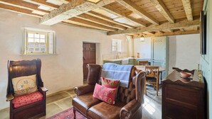 Une pièce rustique avec des poutres apparentes, un fauteuil en cuir agrémenté d’une couverture, une table à manger en bois et, au fond, un coin cuisine.