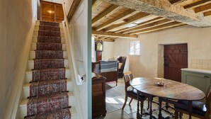 Un escalier recouvert d’une moquette à motifs, une table à manger en bois avec des chaises, et un plafond rustique avec des poutres apparentes.