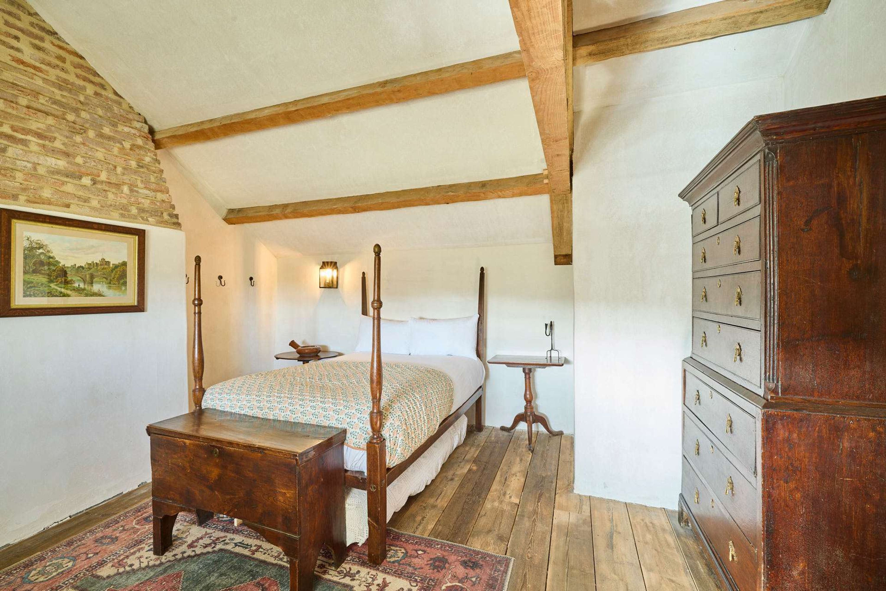 A bedroom with a four-poster bed, wooden dresser, and a small table with a lamp.