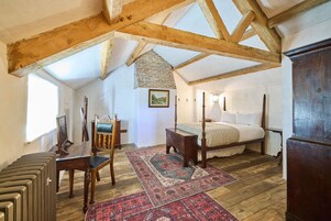 A bedroom with a four-poster bed, wooden beams, a stone fireplace, and a large window.