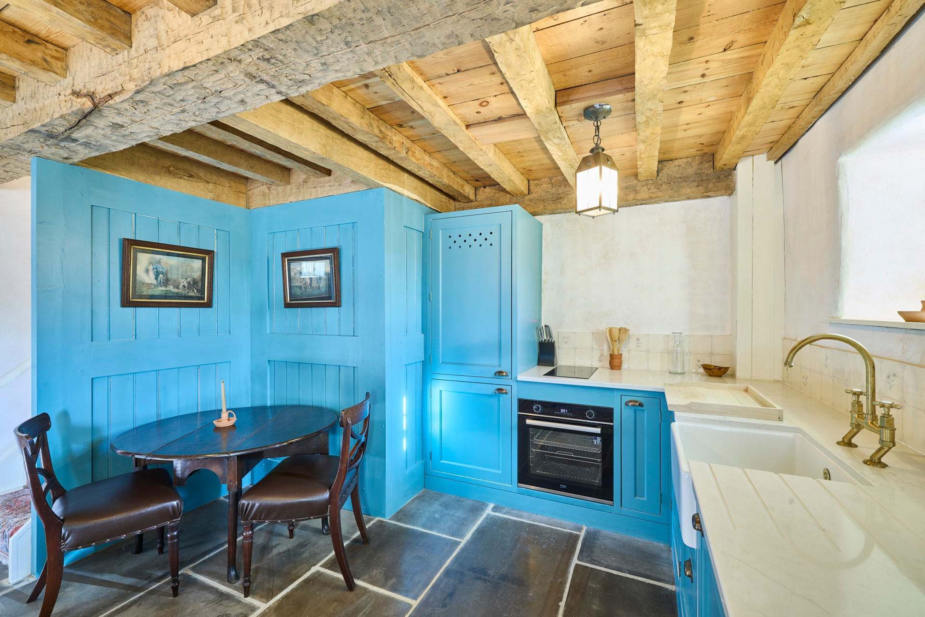 A kitchen with blue cabinets, a wooden dining table with chairs, a built-in oven, and a sink with a brass faucet.