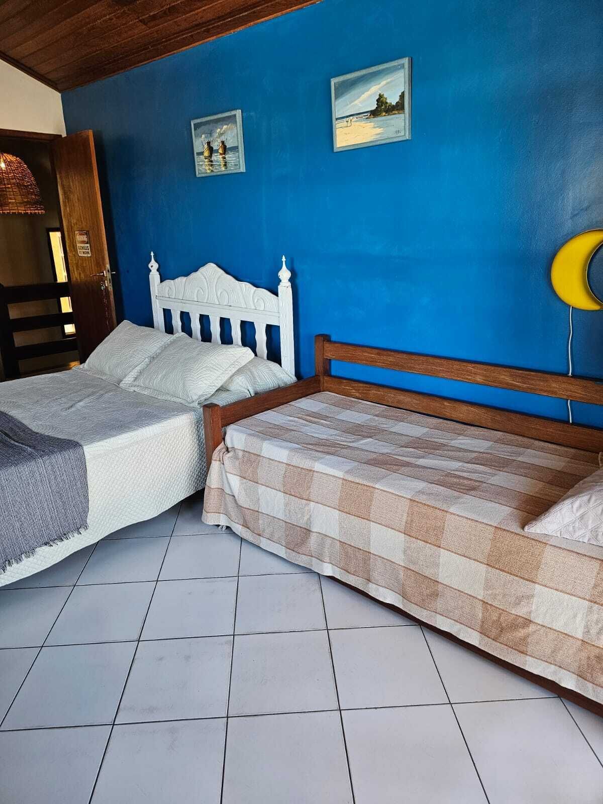 A room with two beds, one with a white headboard and the other with a wooden headboard. There are two framed pictures on the wall, a wooden door, and a yellow object on the right side.