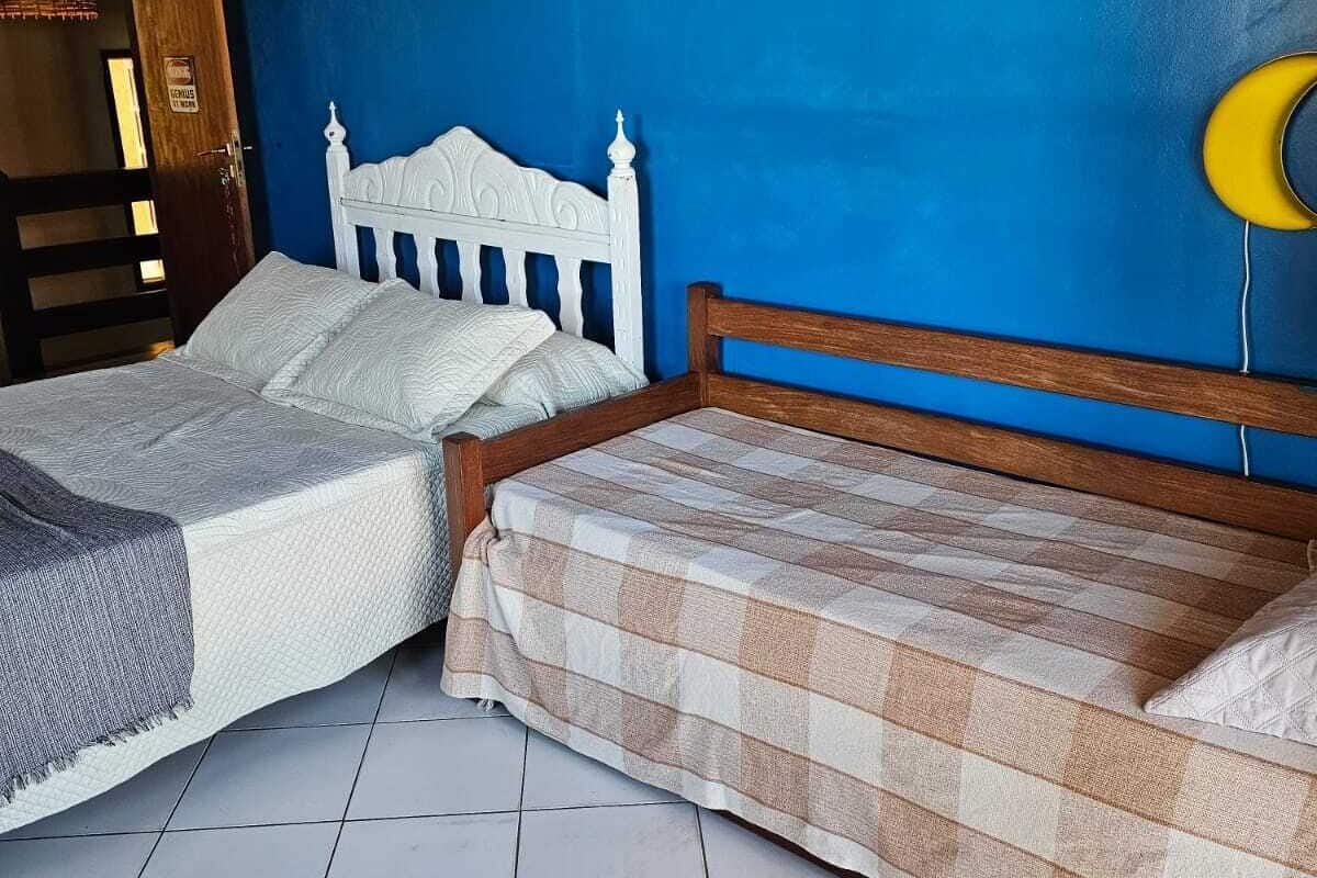Habitación con dos camas, una con cabecera blanca y la otra con cabecera de madera. Se observan dos cuadros en la pared, una puerta de madera y un objeto amarillo a la derecha.