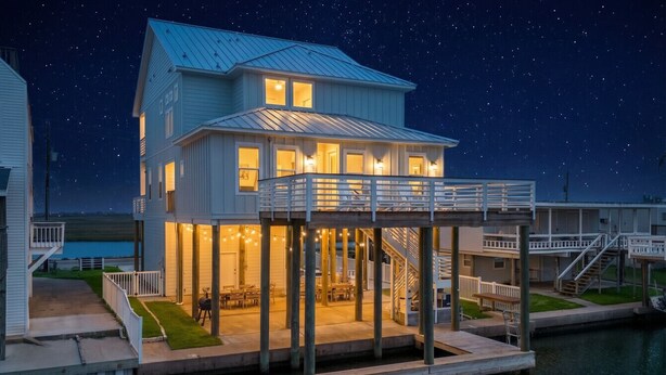 A waterfront house with a covered deck, outdoor seating, and a staircase leading to the upper level.