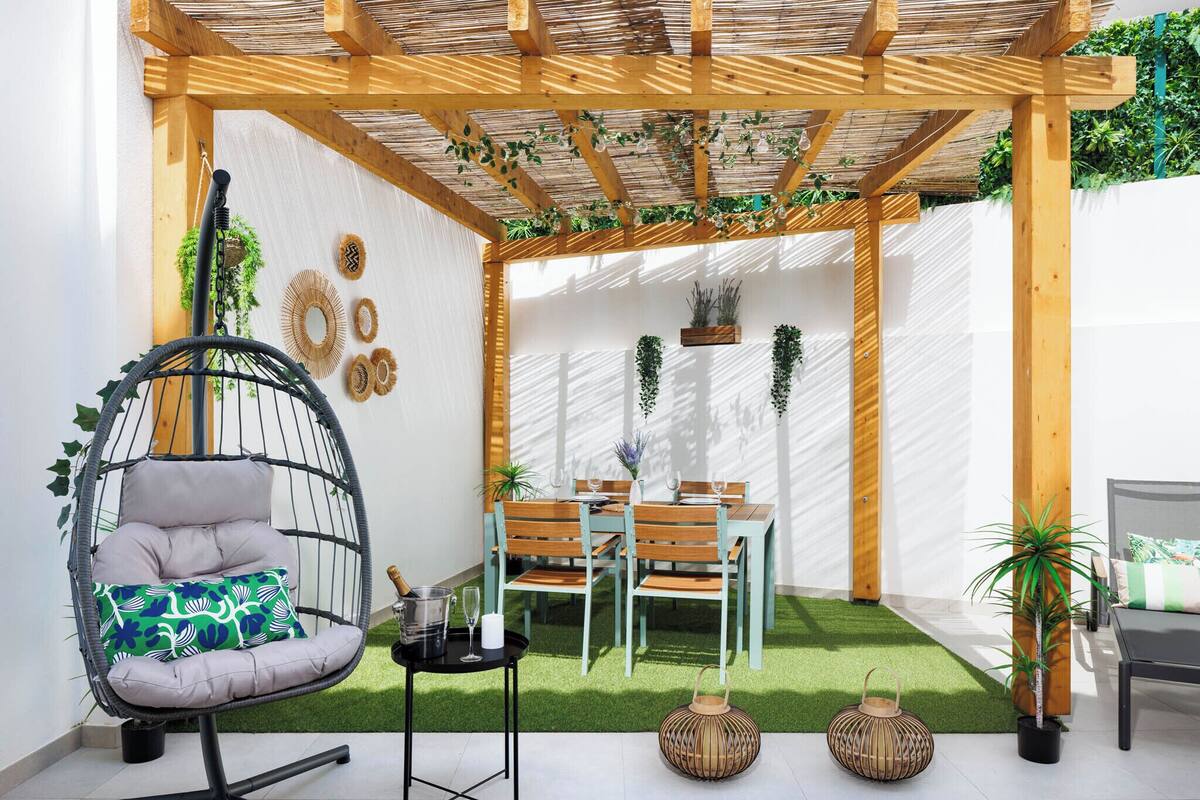Un patio acogedor con una hamaca colgante, una mesa de comedor con sillas y una pequeña zona ajardinada con césped artificial.
