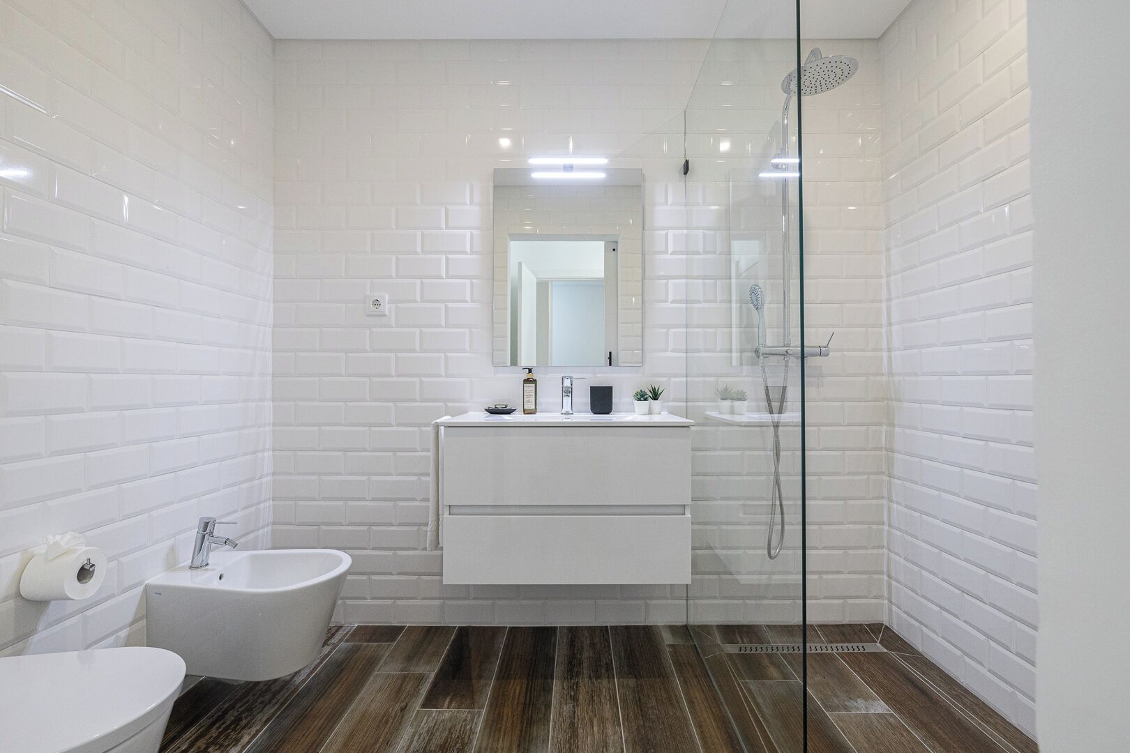 Een moderne badkamer met witte metro tegels, een douche met glazen wand, een bidet en een wastafelmeubel met spiegel.