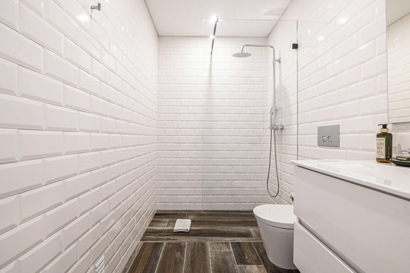 Ein modernes Badezimmer mit weißen Fliesen im U-Bahn-Stil, einer Dusche mit Handbrause, einer Toilette und einem Waschtisch mit einer Shampooflasche.