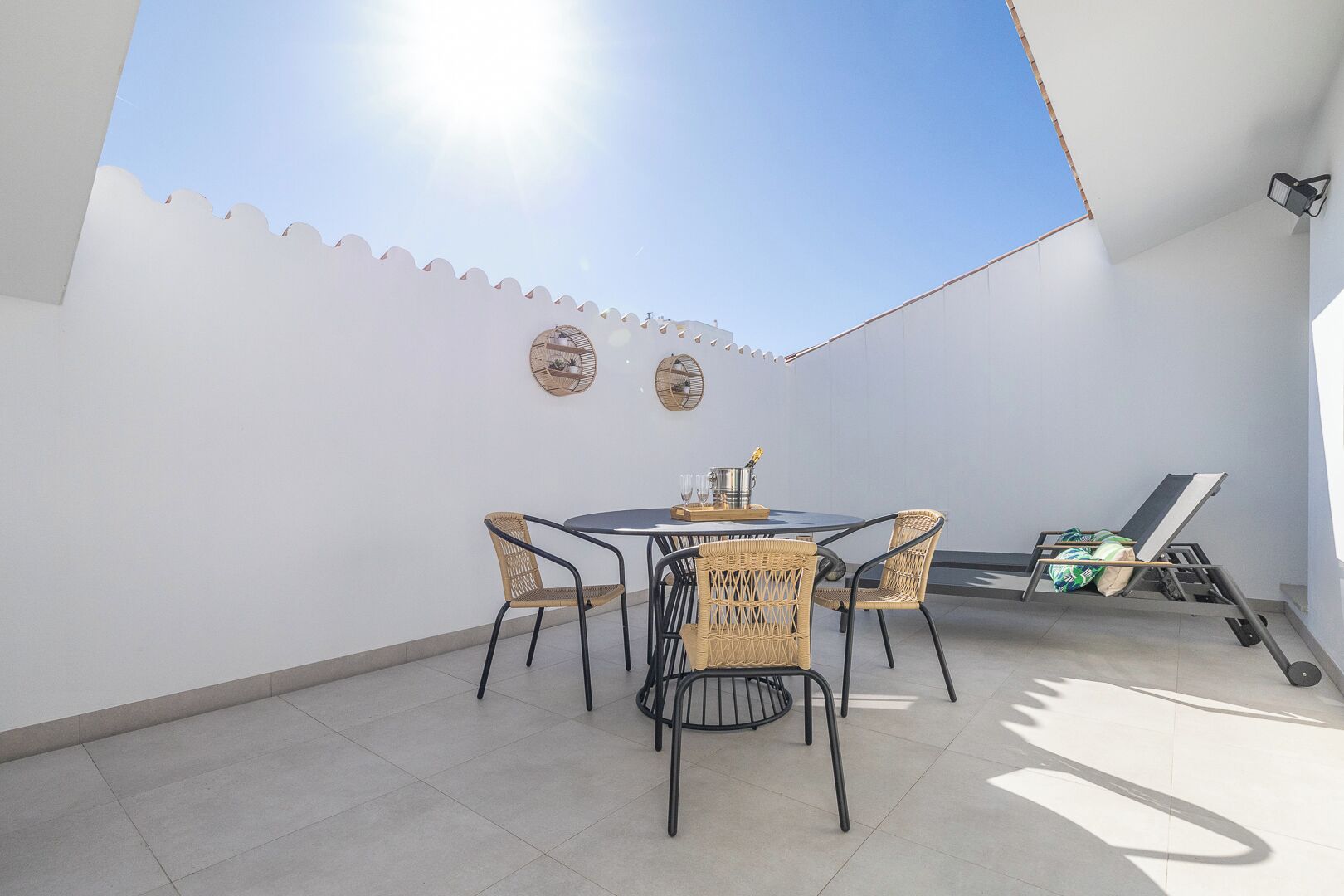 A sunlit patio with a round table and chairs, a lounge chair, and decorative wall art.