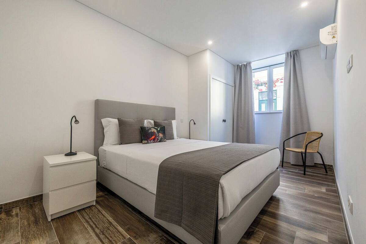 Un dormitorio moderno con cama, mesitas de noche, una silla y un ventanal con cortinas.
