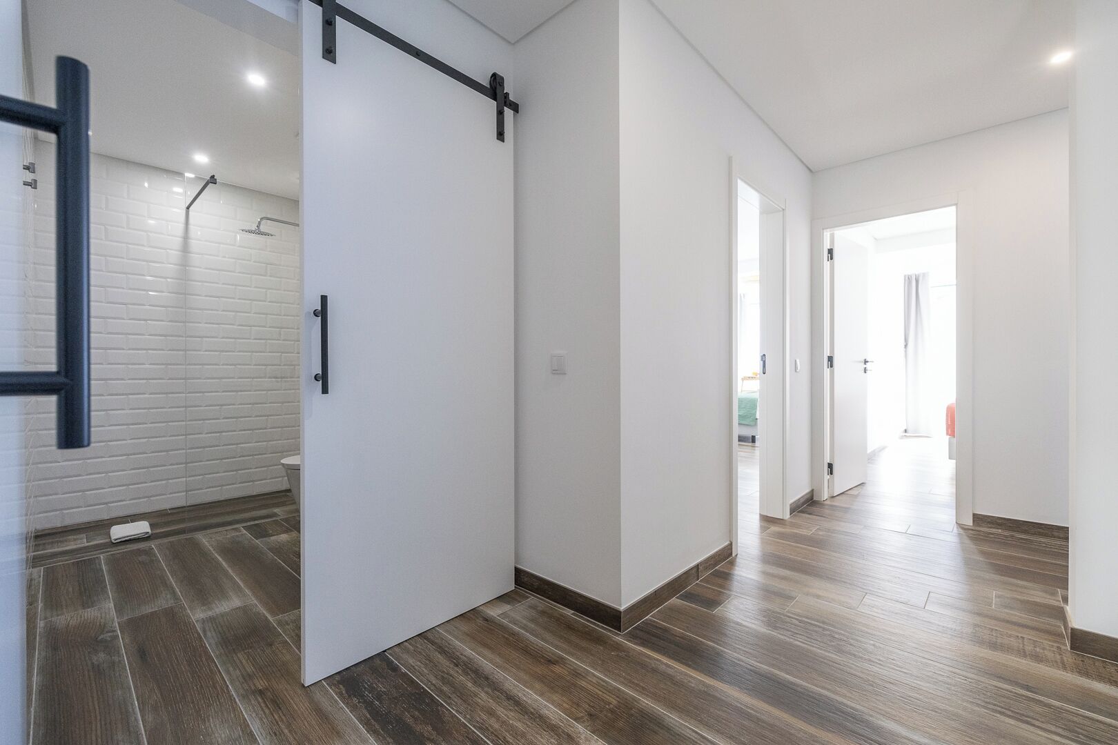 Un interior moderno con una puerta corredera que da al baño, un pasillo con suelo de madera y una habitación luminosa al fondo.