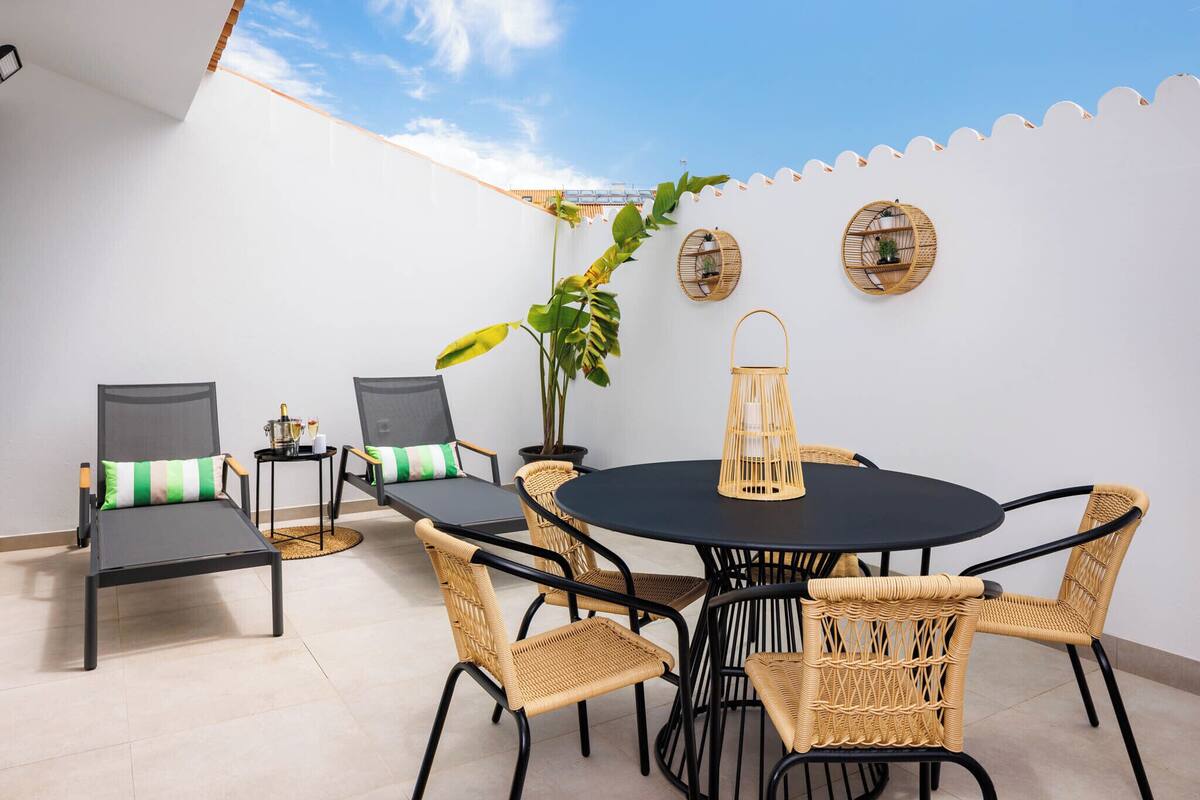 Un patio al aire libre con una mesa redonda y sillas de mimbre, rodeado de paredes blancas y un cielo despejado.