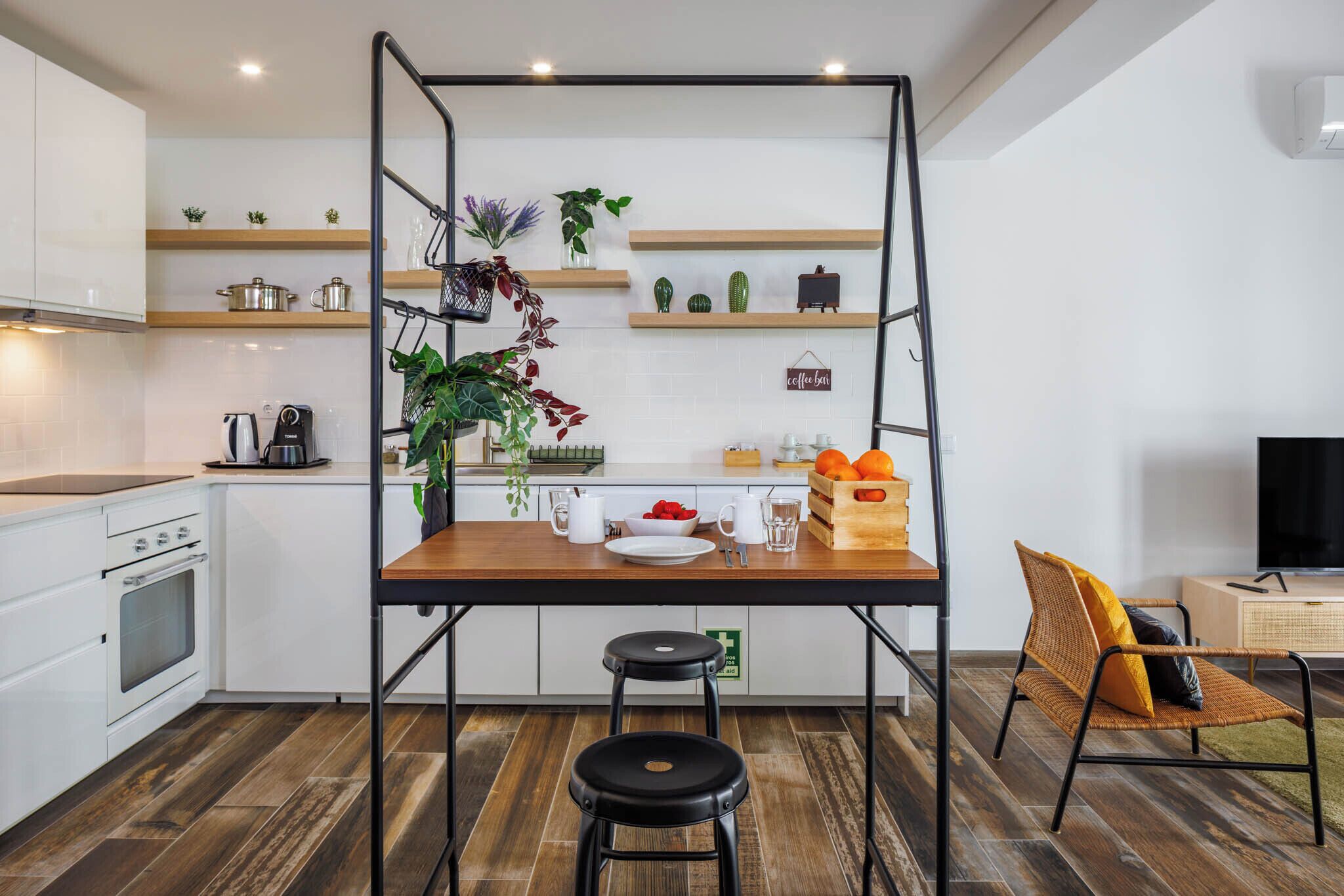 Eine moderne Küche mit weißen Schränken, einer Holzarbeitsplatte und einem schwarzen Metalltisch mit Hocker. Regale mit Pflanzen und Küchenutensilien, eine Kaffeemaschine und ein Fernseher sind im Hintergrund zu sehen.