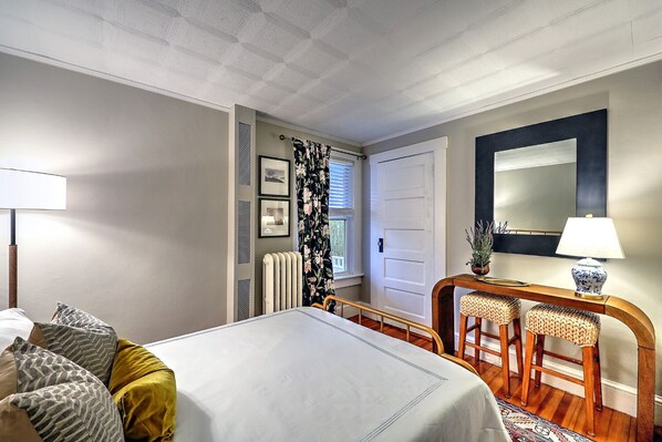 A hotel room with a bed, a desk with a chair, a radiator, a window with curtains, and a mirror on the wall.