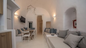 A modern, minimalist living space with a dining area, a sofa with cushions, and a television mounted on the wall. - Cave Villa Valente (Santorini)