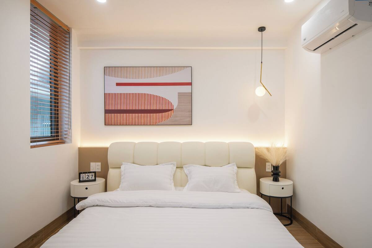 A modern hotel room with a large bed, two bedside tables, a wall-mounted artwork, a ceiling light, and an air conditioning unit.