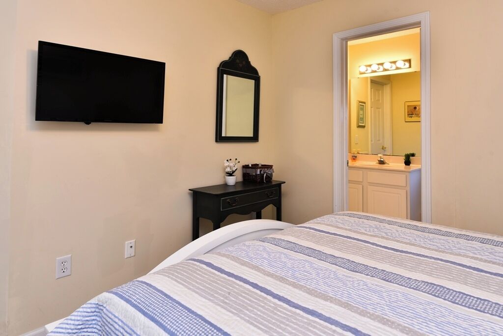 A bedroom with a bed, a flat-screen TV, a small bedside table with a lamp, and a mirror.