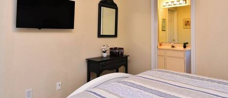 A bedroom with a bed, a flat-screen TV, a small bedside table with a lamp, and a mirror.