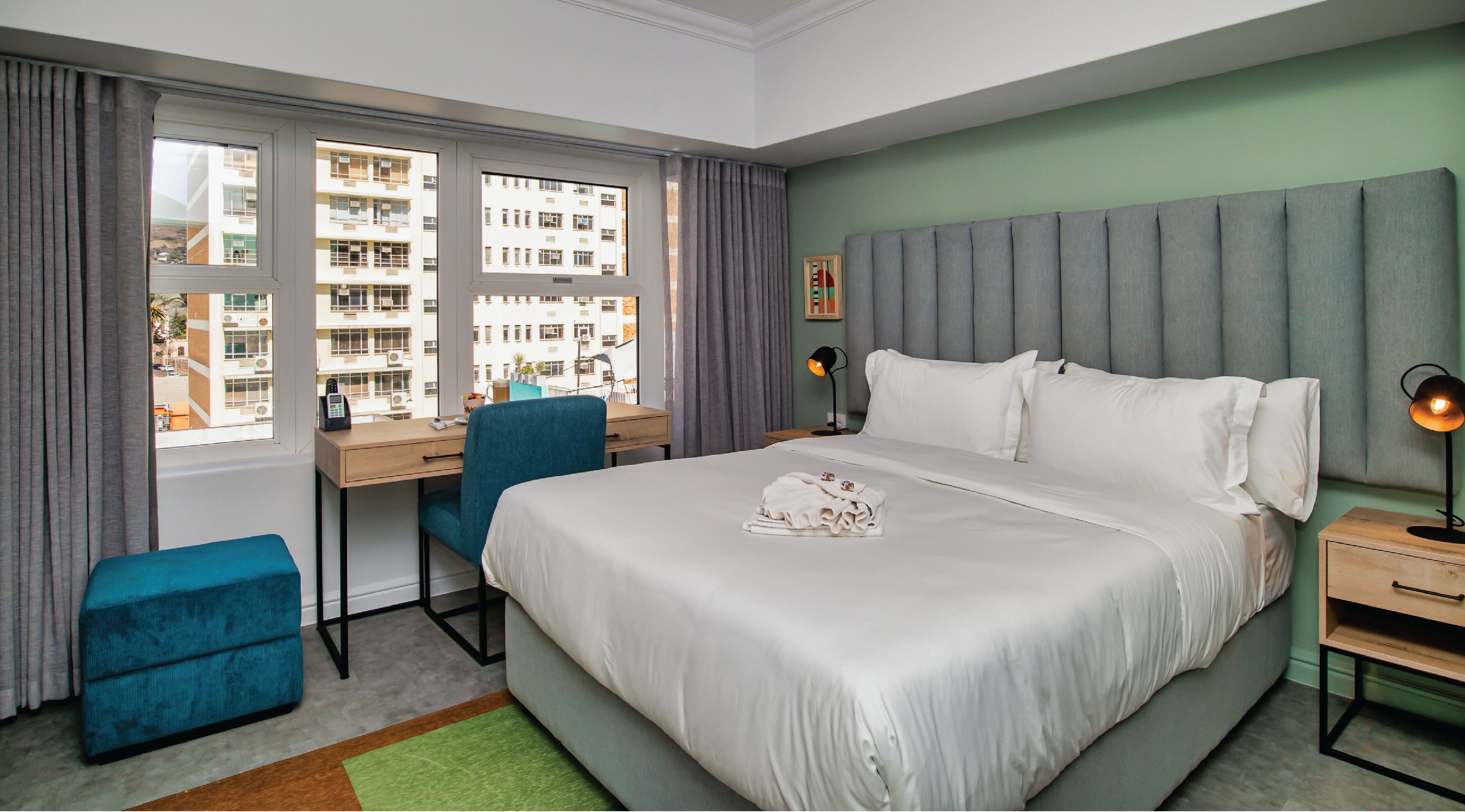 A hotel room with a large bed, a desk, a chair, and a view of the city through the window.