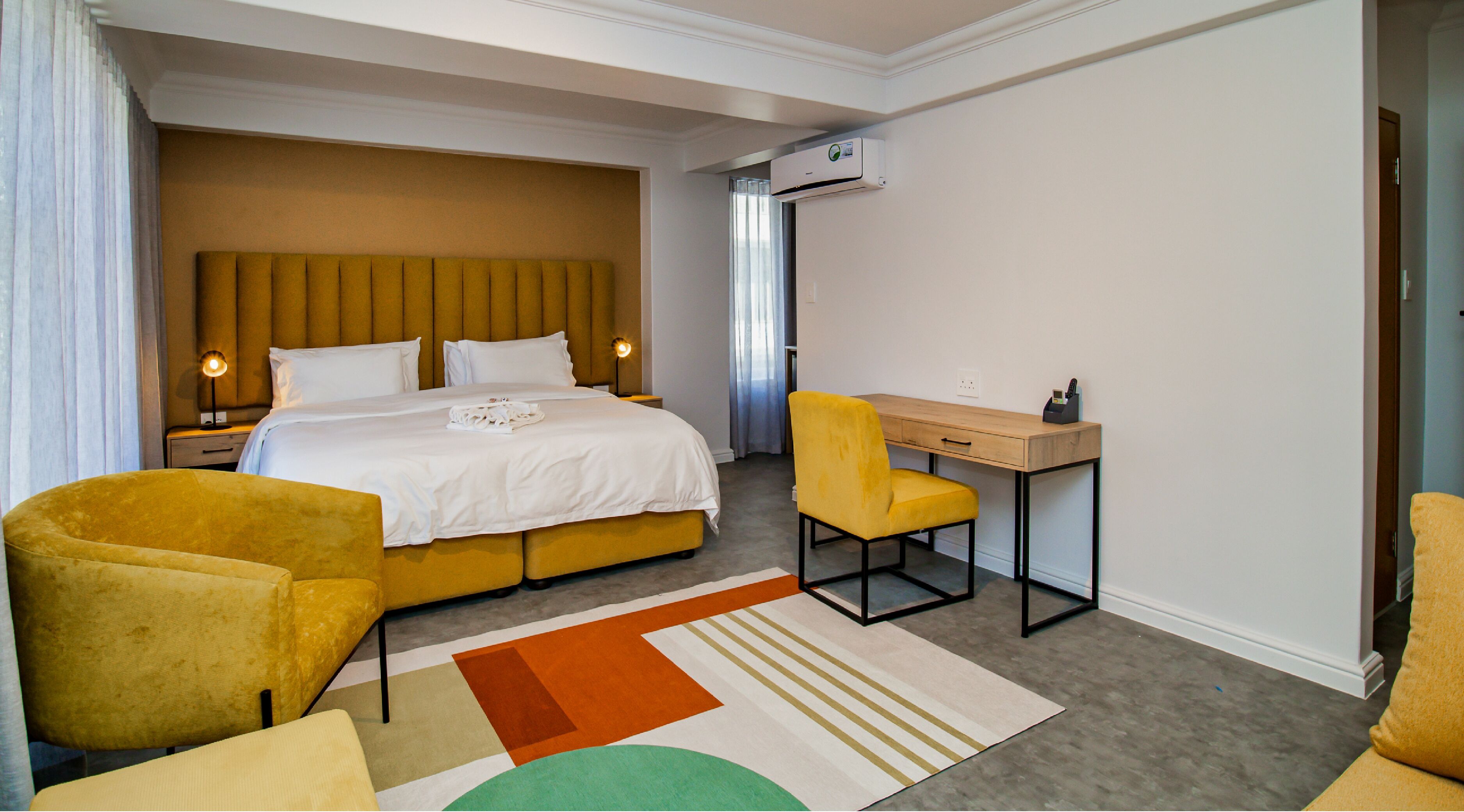A hotel room with a bed, a desk, a chair, a sofa, and an air conditioning unit.