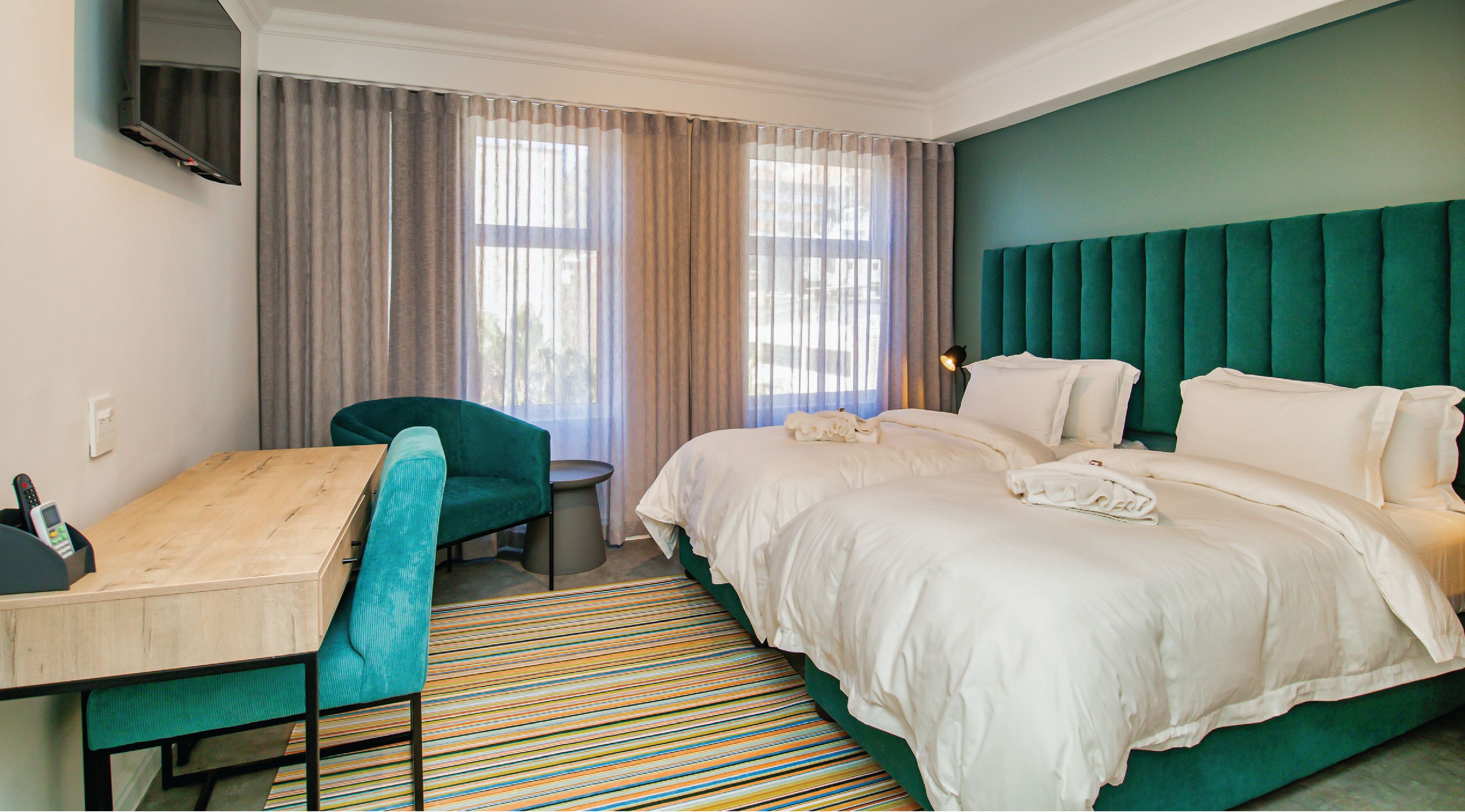 Een hotelkamer met een bed, een bureau, een stoel, een aan de muur bevestigde televisie en een raam met gordijnen.