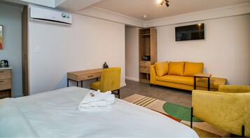 Habitación de hotel con cama, escritorio, sillón amarillo, sofá y televisor montado en la pared.