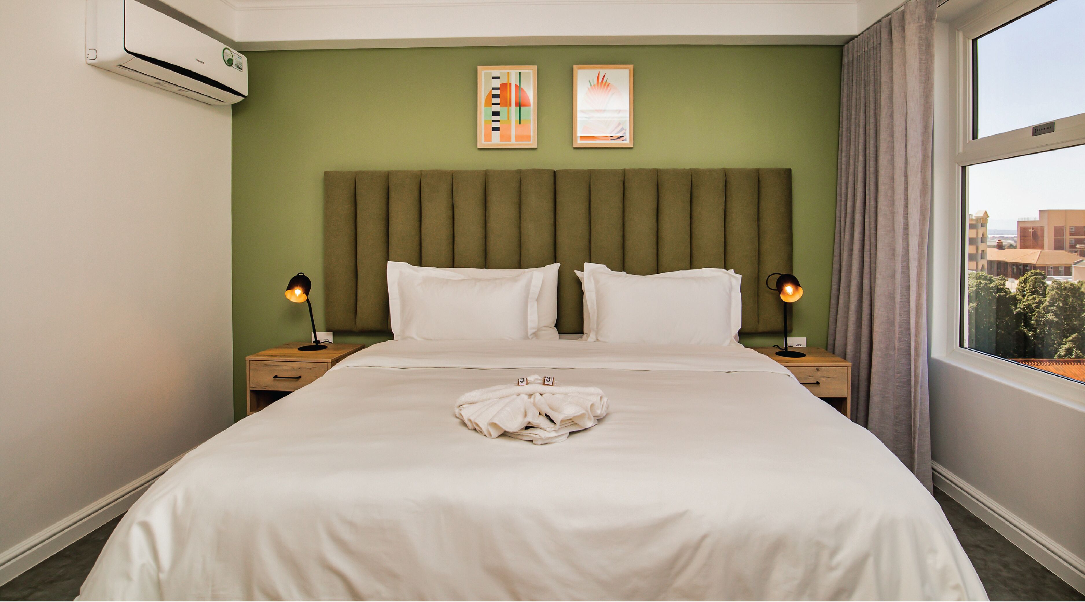 A hotel room with a large bed, two bedside tables, a headboard, two framed artworks, and a window with curtains.