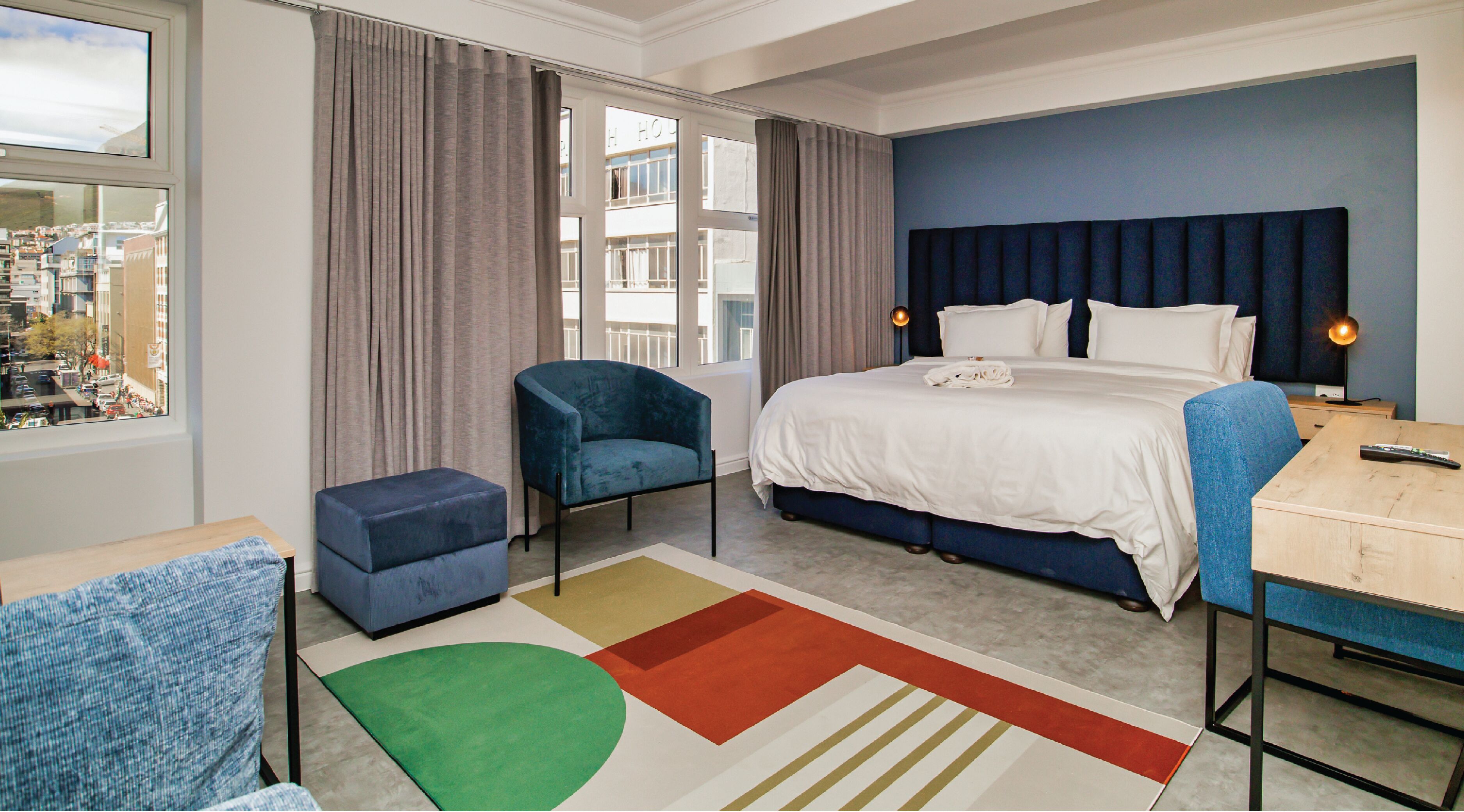 A hotel room with a bed, a chair, a desk, and a view of the city.