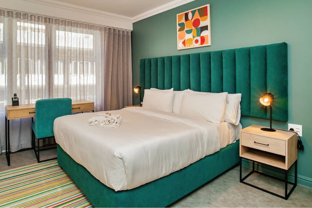 A hotel room with a large bed, a desk, a chair, and a colorful artwork on the wall.