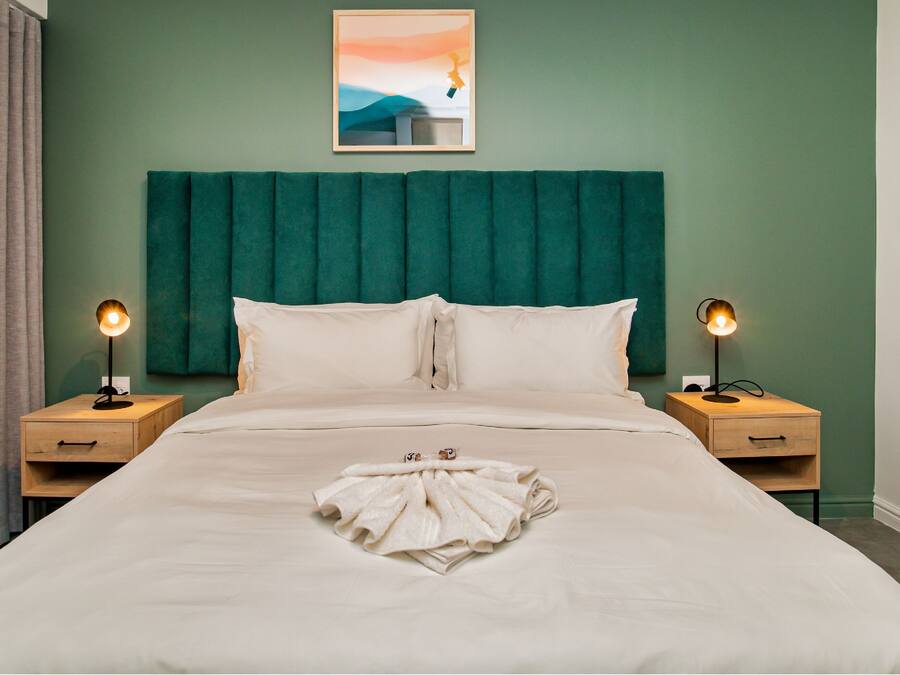 Quarto de hotel com uma cama grande, duas mesas de cabeceira com luminárias, cabeceira verde, um quadro na parede e vista para o exterior.