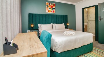A hotel room with a bed, bedside tables, a desk, a shower area, and a colorful abstract painting on the wall.