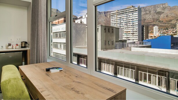 A hotel room with a wooden table, a sofa, and large windows offering a view of a cityscape and mountains. - 9 On Barrack (Cape Town)
