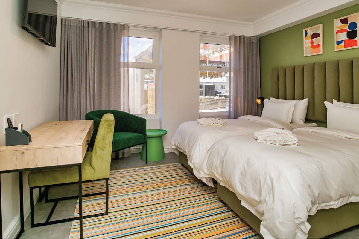 A hotel room with a bed, a desk, a chair, a green stool, a striped rug, and artwork on the wall.