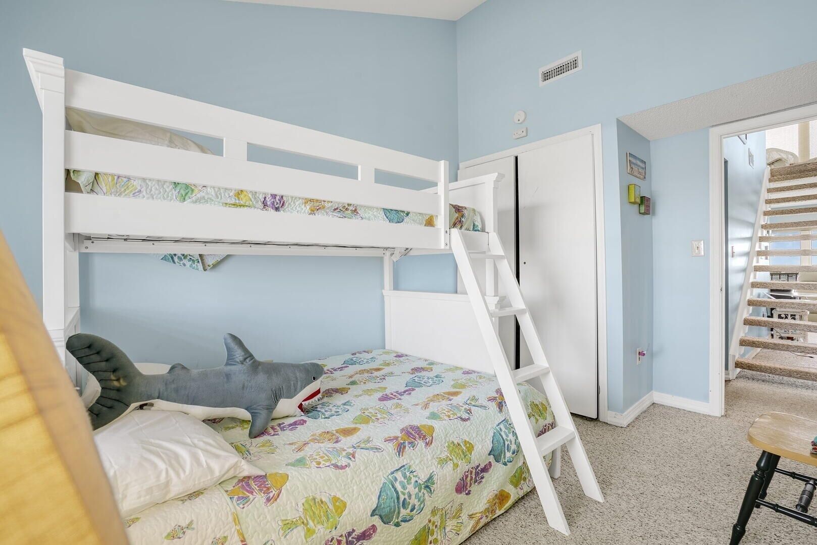 A bunk bed room with a ladder, a bed with a colorful fish-patterned bedspread, and a door leading to another room.