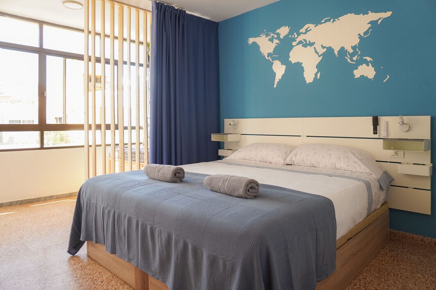 Une chambre d’hôtel avec un grand lit, une carte du monde accrochée au mur, une fenêtre avec des stores et un rideau.
