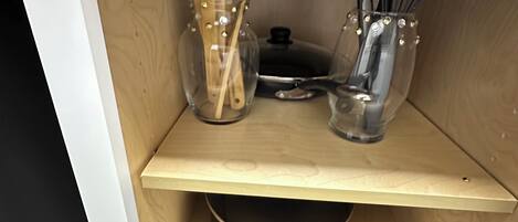 Ein Küchenschrank mit einer Holzwand, zwei Glasbehältern mit Metalldeckeln, einem Metalltopf und Küchenutensilien aus Holz und Metall.