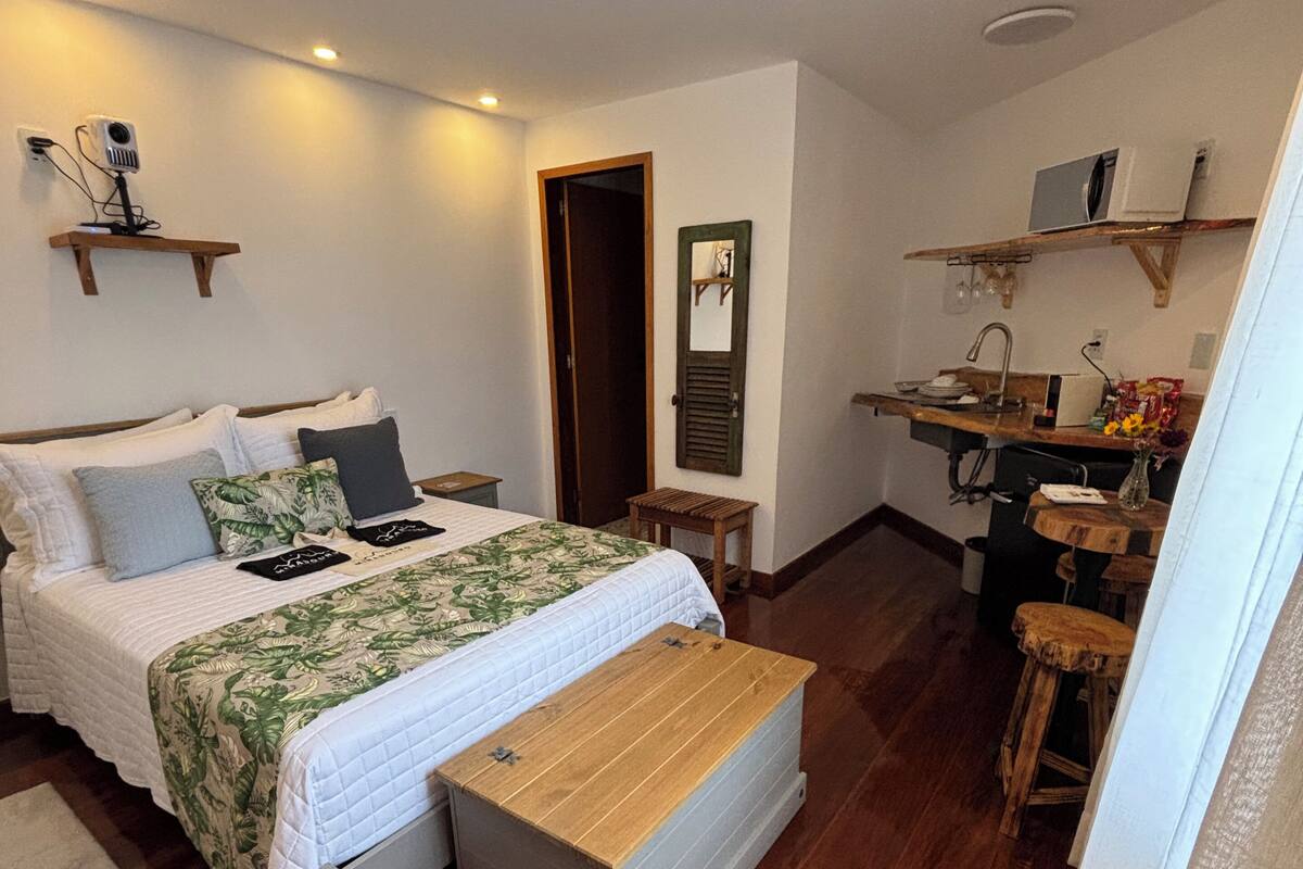 A bedroom with a bed, bedside tables, a wooden bench, a small stool, and a kitchenette area with a sink and a microwave.