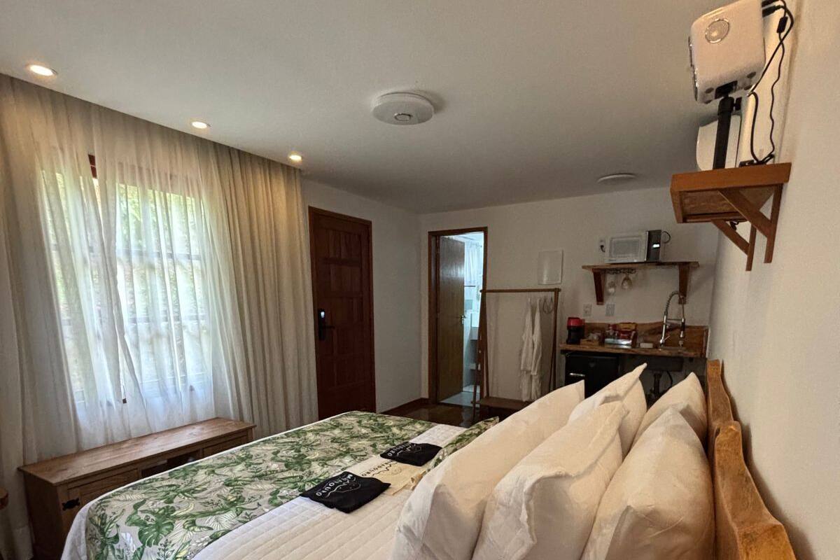 A bedroom with a bed, a wooden bench, a desk, and a kitchenette in the background.