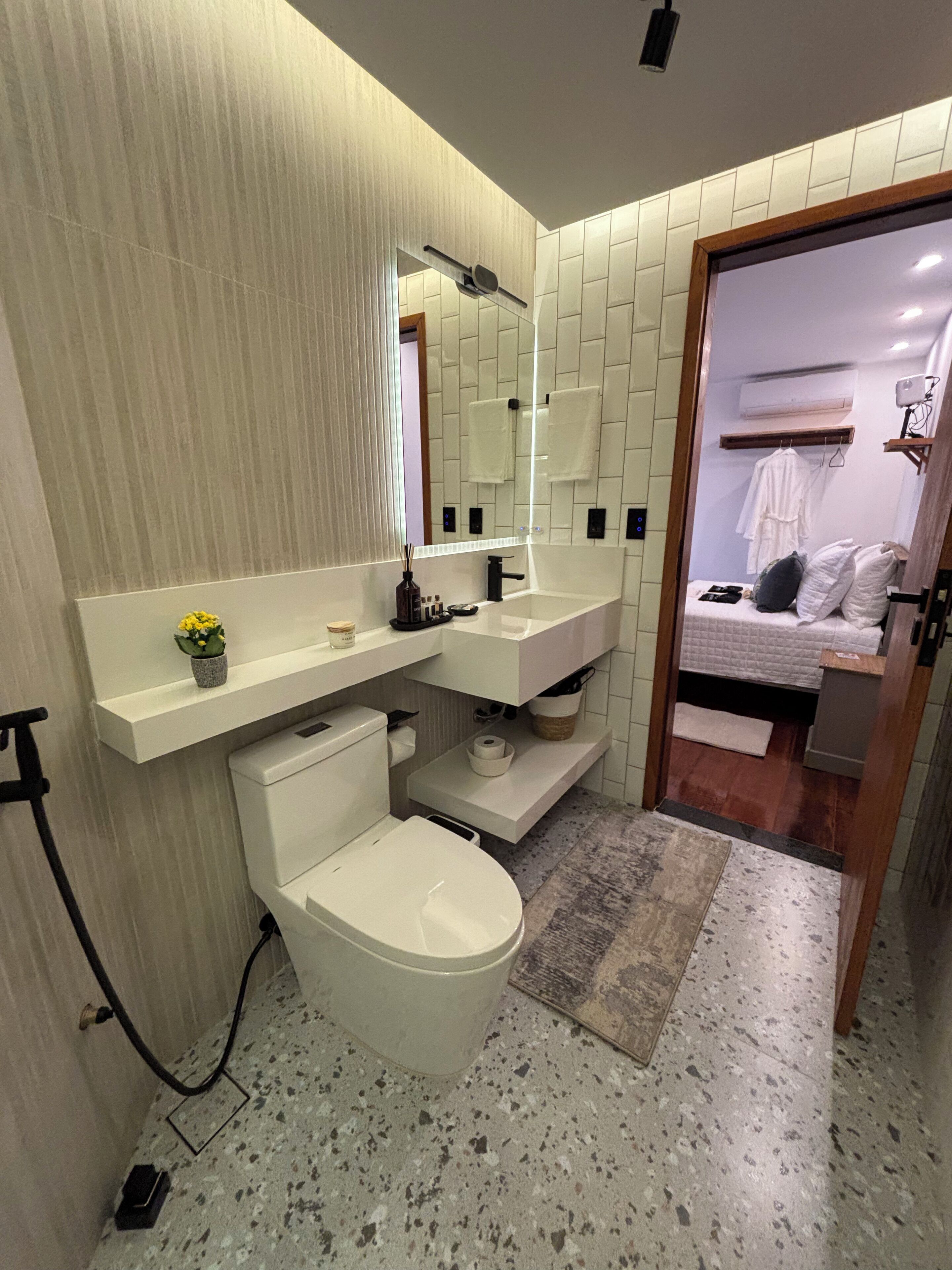 A compact bathroom with a toilet, sink, and mirror. There is a small shelf with toiletries and a potted plant. The floor is speckled, and the walls are tiled.