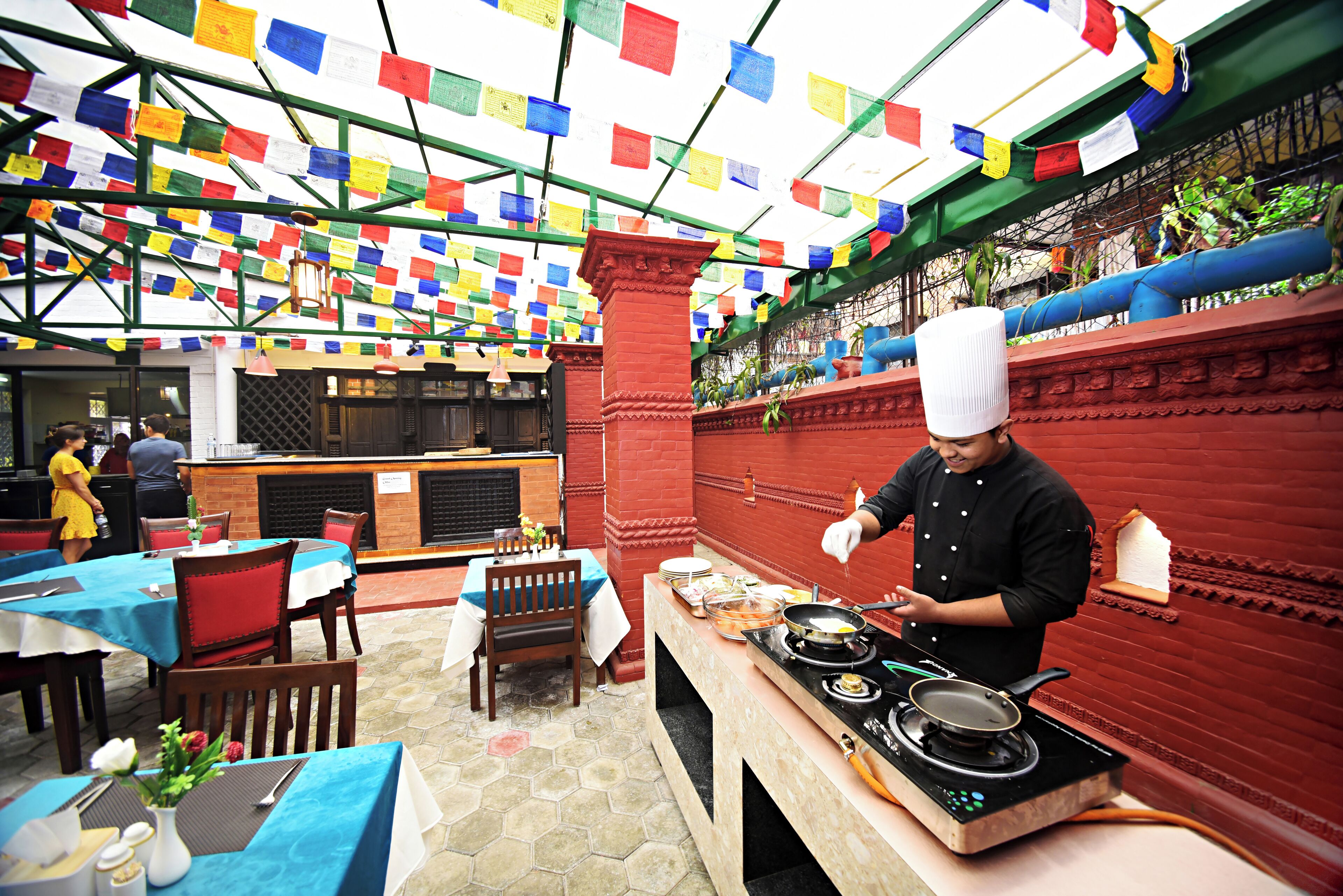 A chef cooking in a restaurant with colorful prayer flags, tables, and chairs.